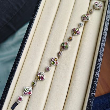 A delicate silver-toned bracelet with floral clusters of pink, green, and clear stones, displayed inside an open cream-colored jewelry box.