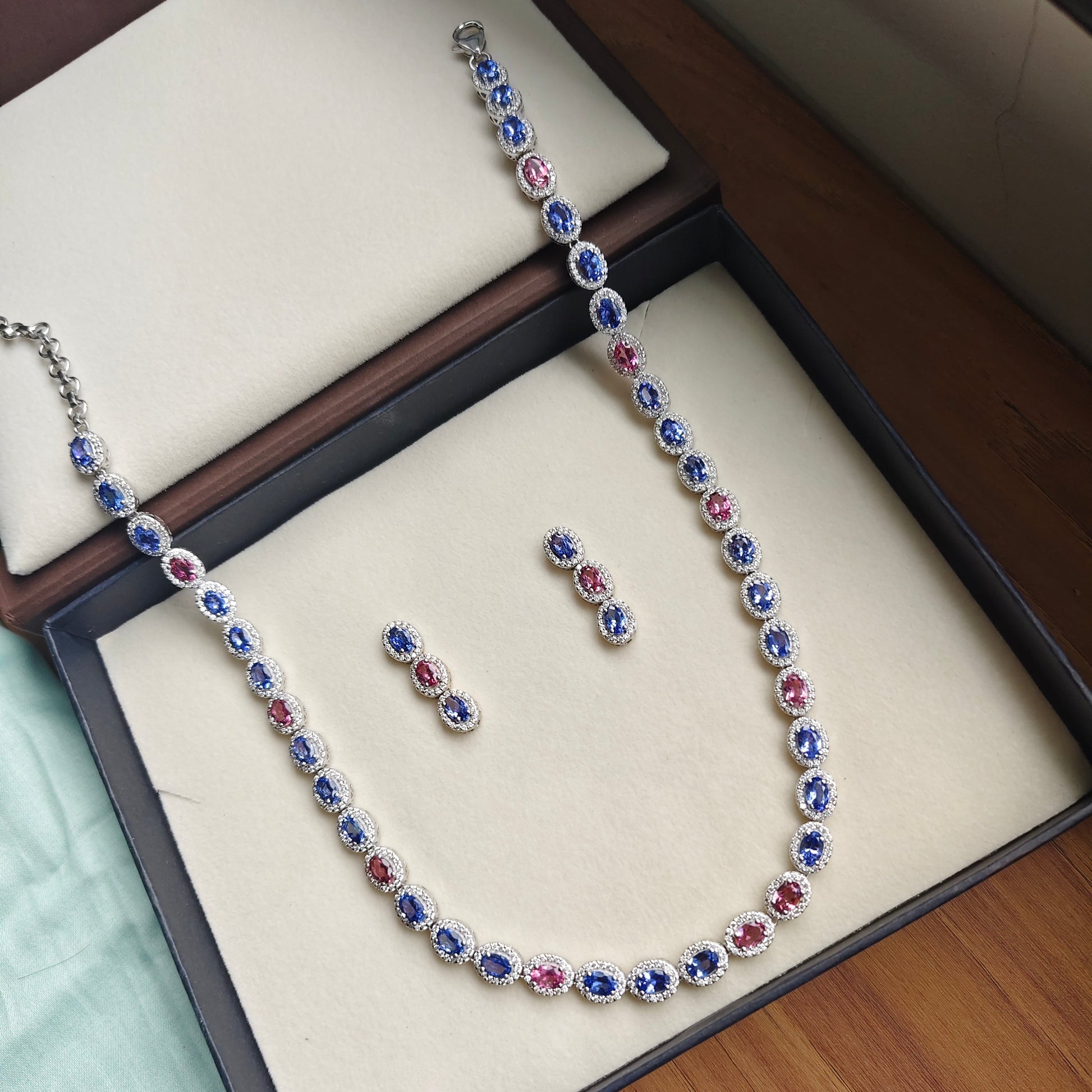 A silver-toned necklace and matching earrings set with oval-cut blue, pink, and red gemstones, displayed inside an open jewelry box on a wooden surface.