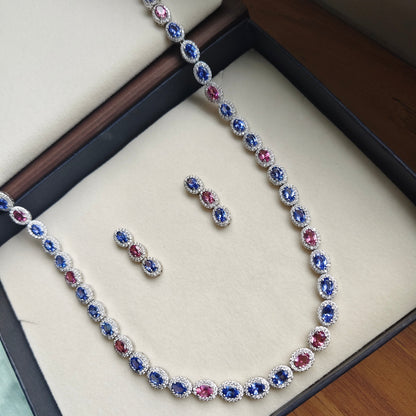 A silver-toned necklace and matching earrings set with oval blue and pink gemstones, displayed inside an open jewelry box on a wooden surface.