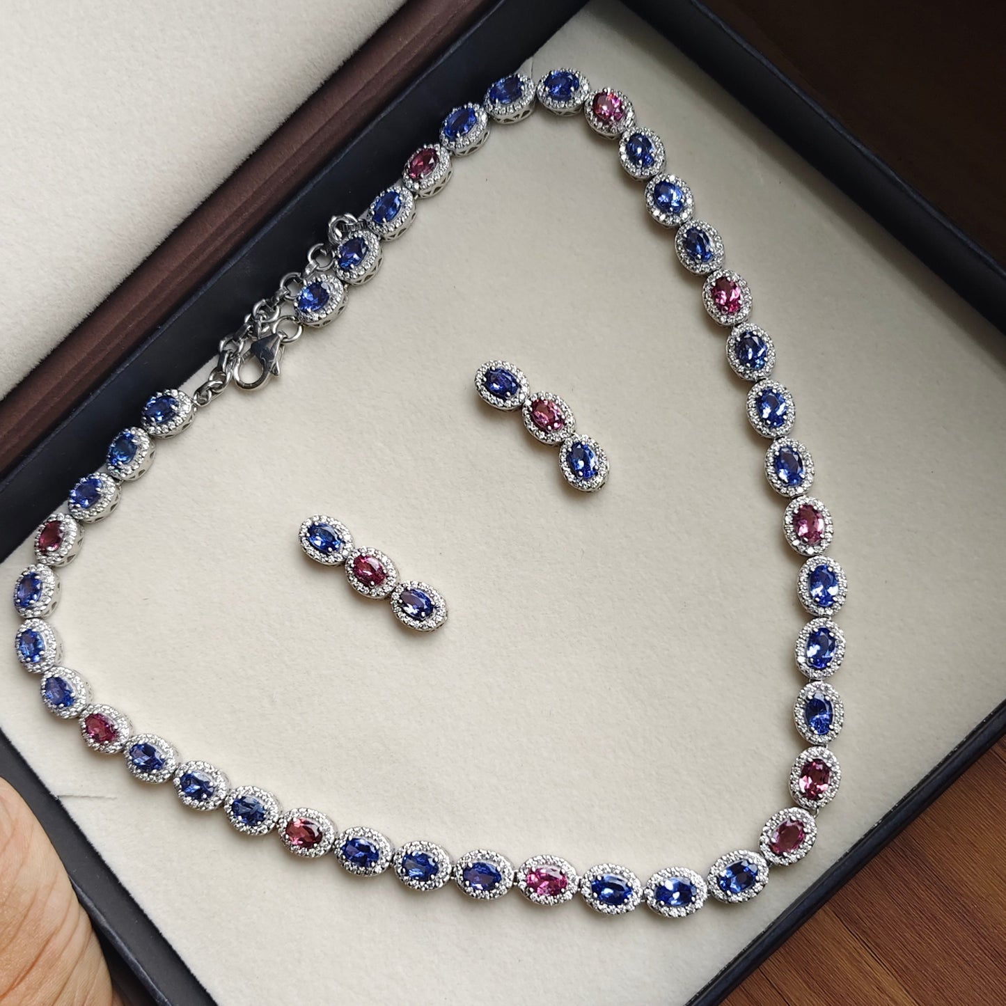 A silver-toned tennis bracelet and matching stud earrings displayed on a cream jewelry box. The jewelry features oval-cut blue and pink stones surrounded by small clear stones in a halo setting.