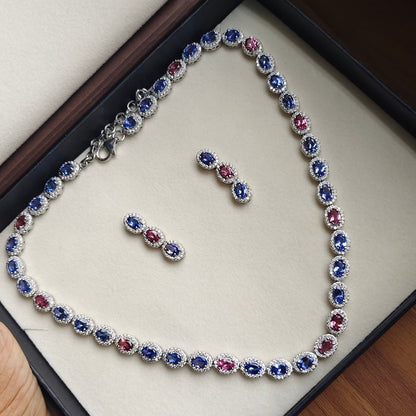 A silver-toned tennis bracelet and matching stud earrings displayed on a cream jewelry box. The jewelry features oval-cut blue and pink stones surrounded by small clear stones in a halo setting.