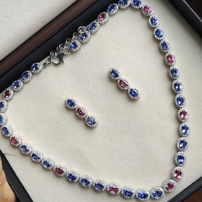 A silver-toned jewelry set featuring a tennis necklace and matching earrings, decorated with alternating oval-cut blue and pink gemstones surrounded by small clear stones, displayed inside a cream-colored jewelry box.