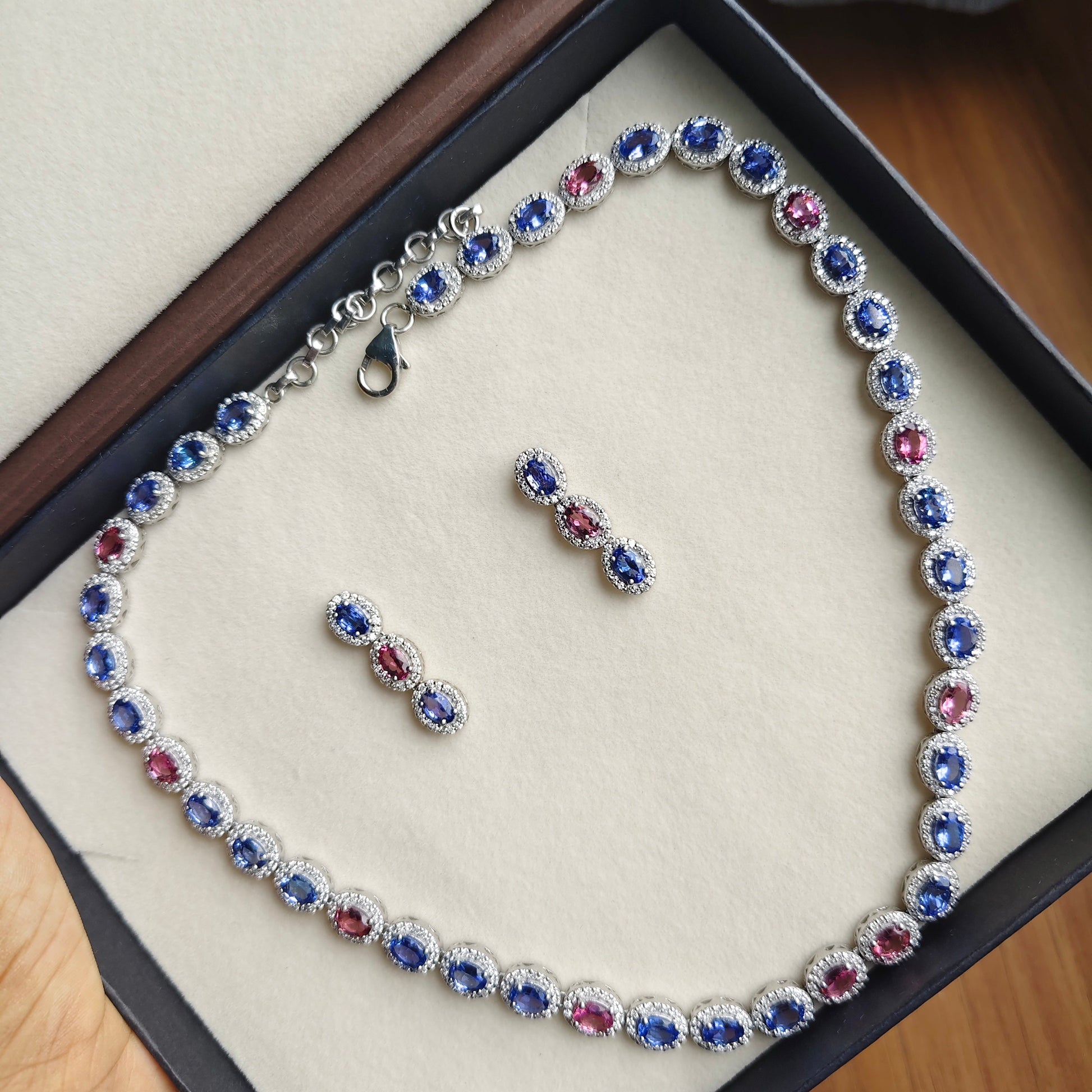 A silver-toned jewelry set with a necklace and matching earrings, featuring round blue and red gemstones surrounded by small clear stones, displayed inside a black jewelry box with a beige insert.