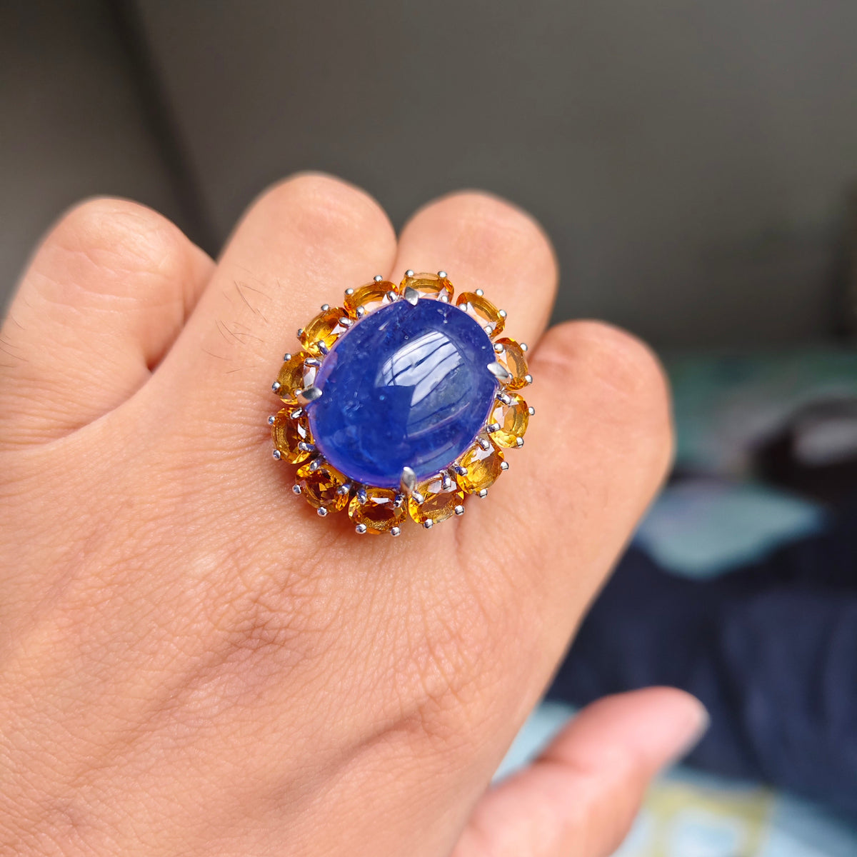 A close-up of a hand wearing a large ring with an oval deep blue cabochon stone surrounded by a halo of small round gold and amber-colored stones.