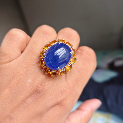 A close-up of a hand wearing a large ring with an oval deep blue cabochon stone surrounded by a halo of small round gold and amber-colored stones.