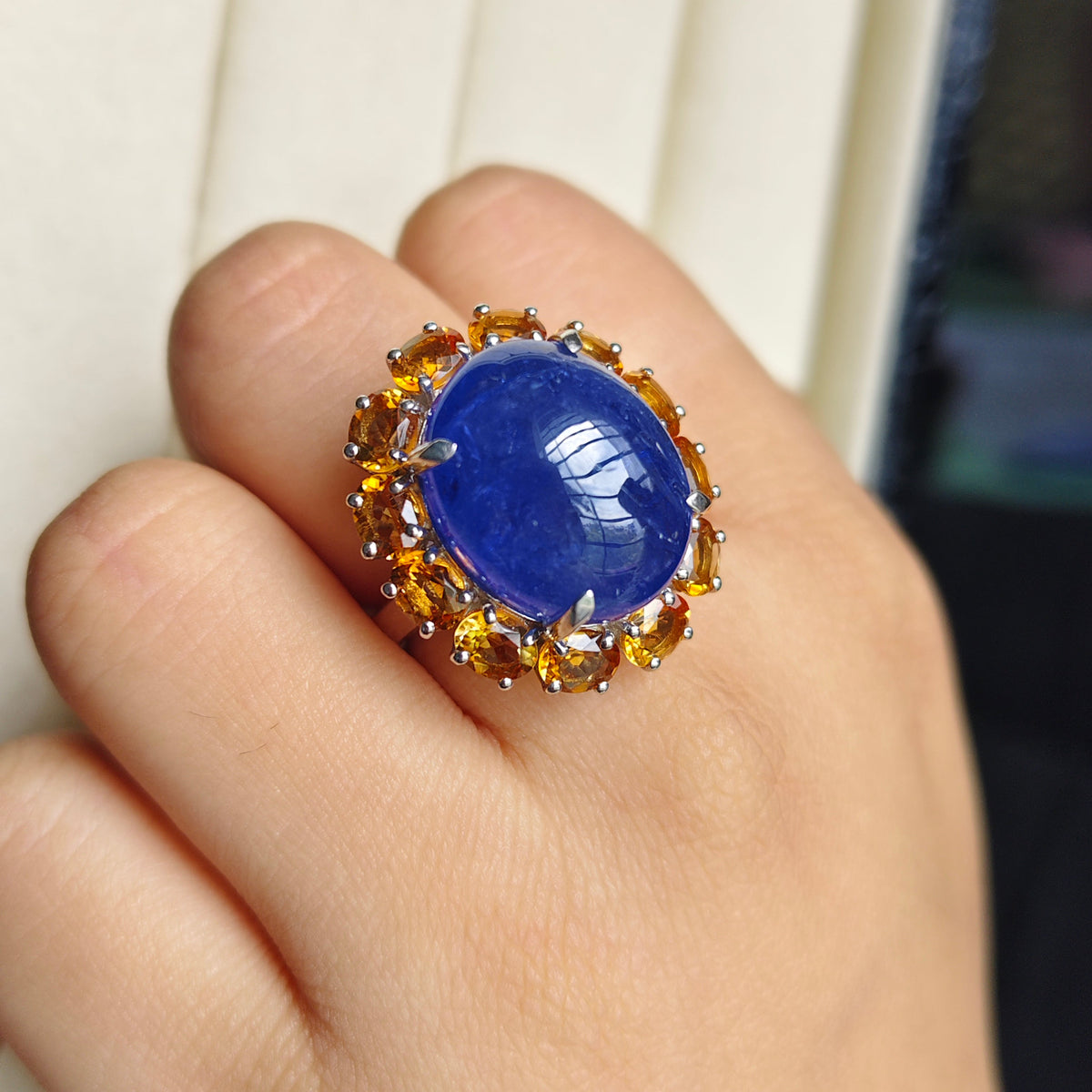 A close-up of a hand wearing a ring with a large oval blue cabochon stone surrounded by small round gold stones.