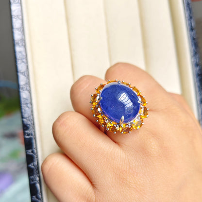 A close-up of a hand wearing a large ring with a round blue cabochon stone surrounded by small gold and silver stones, set against a cream jewelry box.