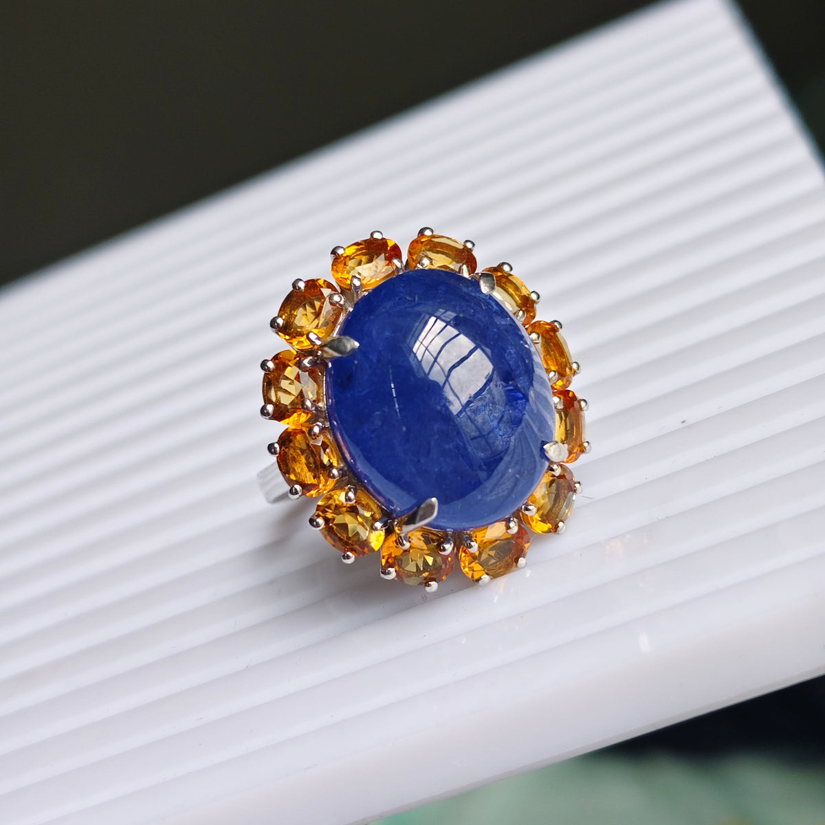 A ring featuring a large oval blue sapphire stone surrounded by a halo of round yellow citrine gemstones in a silver setting, resting on a white ribbed surface.
