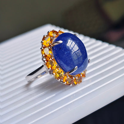 A silver ring featuring a large oval blue lapis lazuli stone surrounded by a halo of round yellow citrine gemstones, resting on a white ribbed surface.
