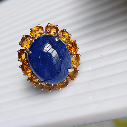 A close-up shot of a ring featuring a large oval cabochon blue sapphire surrounded by a halo of small round-cut yellow citrine gemstones in a silver setting, resting on a white ribbed surface.