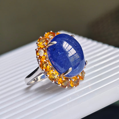 A silver ring featuring a large oval blue lapis lazuli stone surrounded by a halo of round orange citrine gemstones, resting on a white ribbed surface.