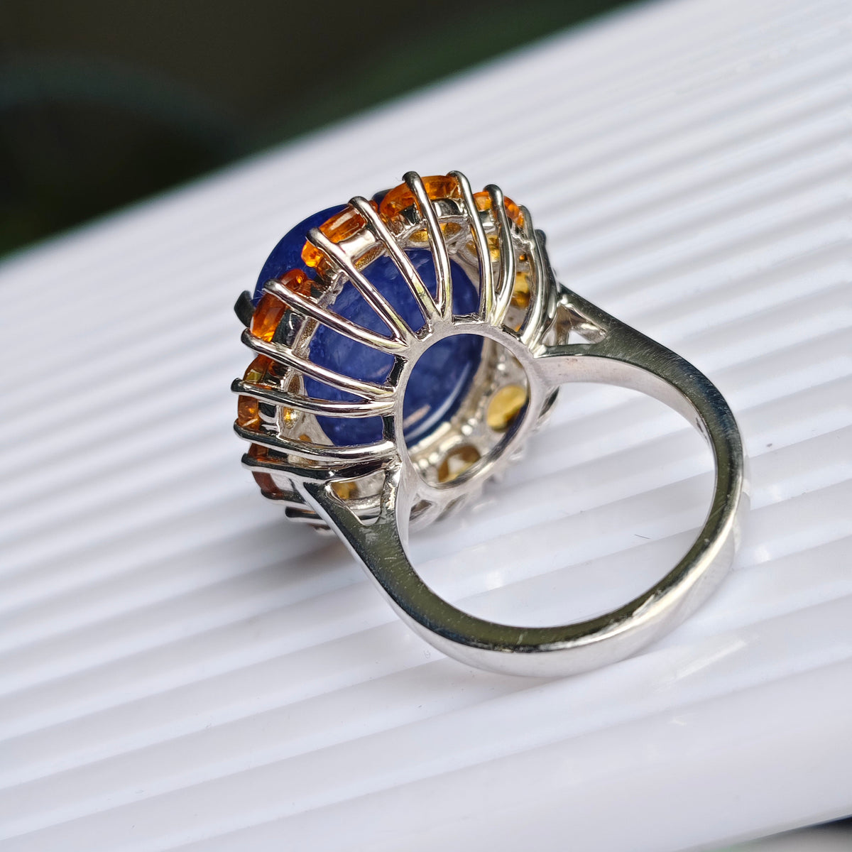 A silver ring featuring a round blue lapis lazuli stone surrounded by a halo of small orange gemstones, resting on a white ribbed surface.