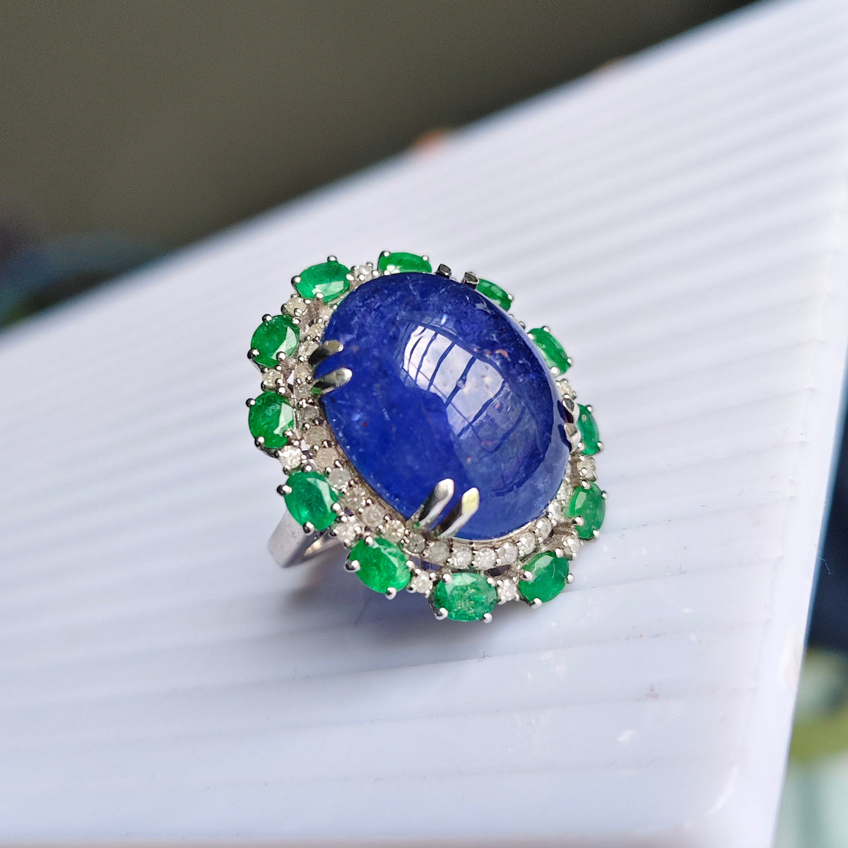 A ring with a large oval blue lapis lazuli stone surrounded by green emerald-cut accents and small clear stones in a silver-toned setting, held in a hand over a white ribbed surface.