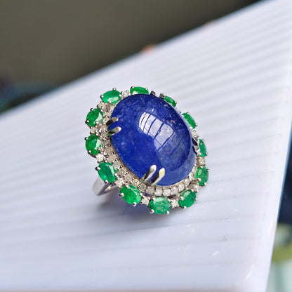 A ring with a large oval blue lapis lazuli stone surrounded by green emerald-cut accents and small clear stones in a silver-toned setting, held in a hand over a white ribbed surface.