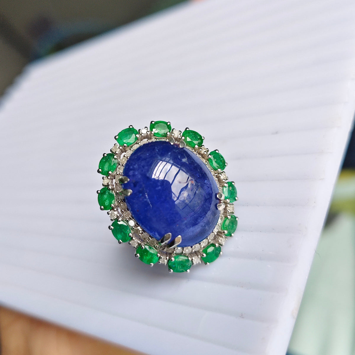 A ring featuring a large oval blue sapphire center stone surrounded by a halo of green emerald-cut gemstones and small clear stones, held on a white ribbed display card.