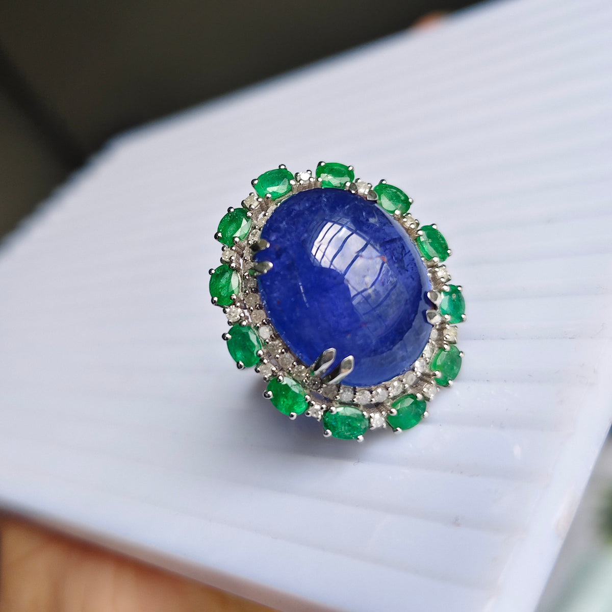 A ring with a large oval blue cabochon stone surrounded by a halo of green emerald-cut stones and small clear accents, held on a white display card.