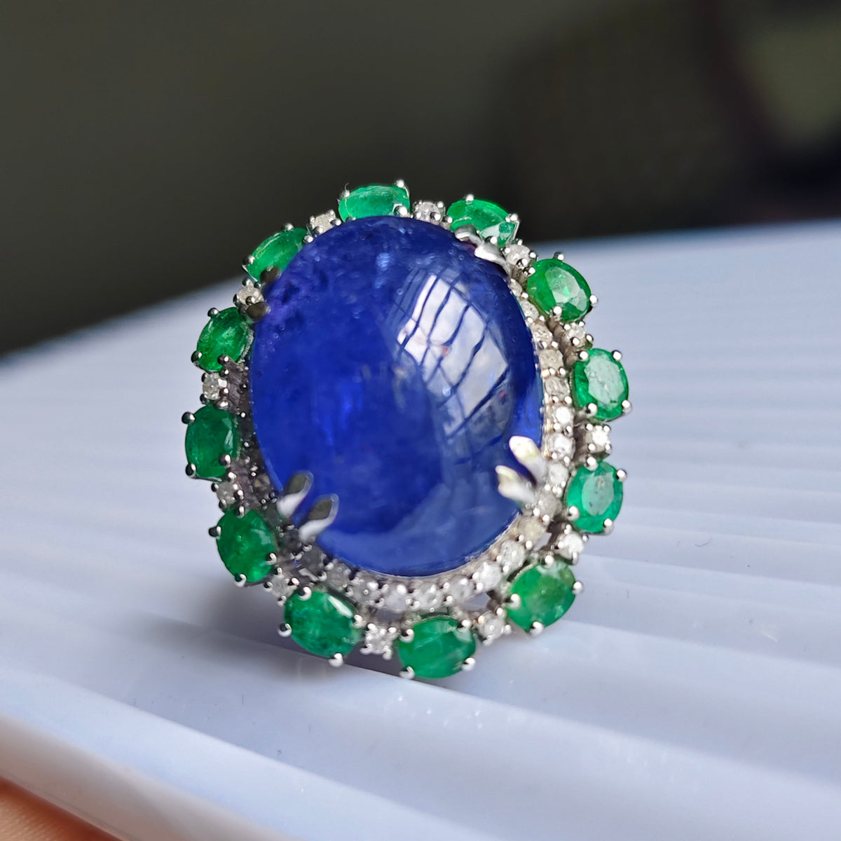 A close-up shot of a ring featuring a large oval blue sapphire center stone surrounded by a halo of green emeralds and small clear stones, resting on a white ribbed surface.