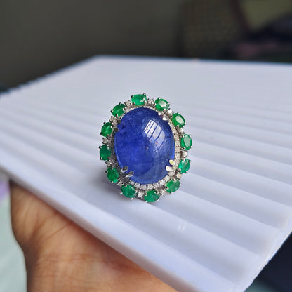 A ring with a large oval blue sapphire center stone and a halo of green emerald-cut side stones, held on a white ribbed display block by a hand.