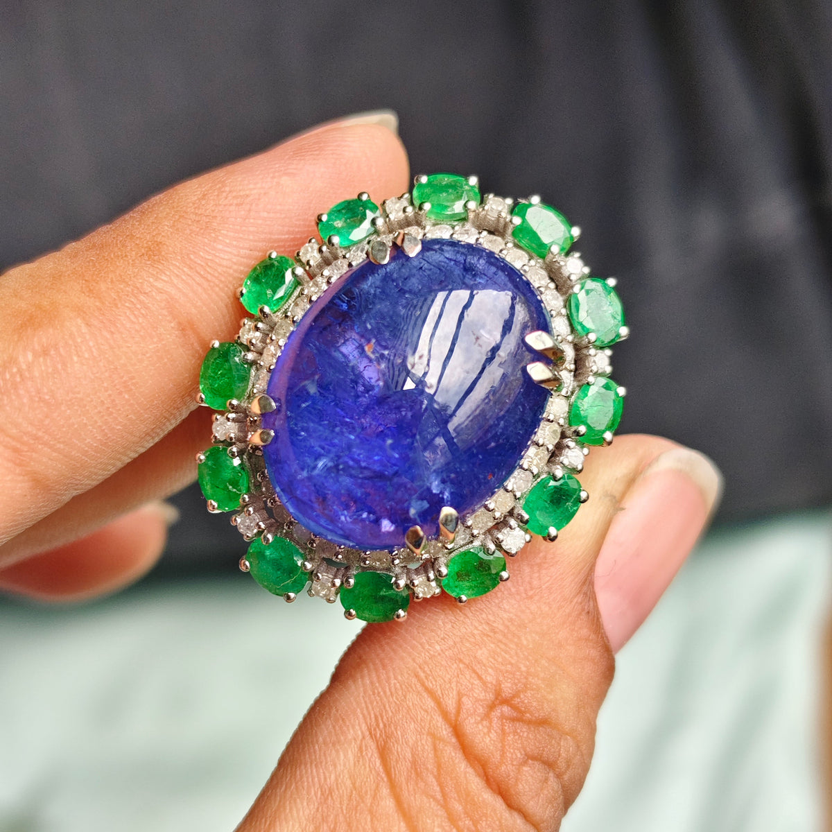 A close-up of a hand holding a large oval ring featuring a deep blue lapis lazuli stone surrounded by a halo of green emeralds and small clear stones.