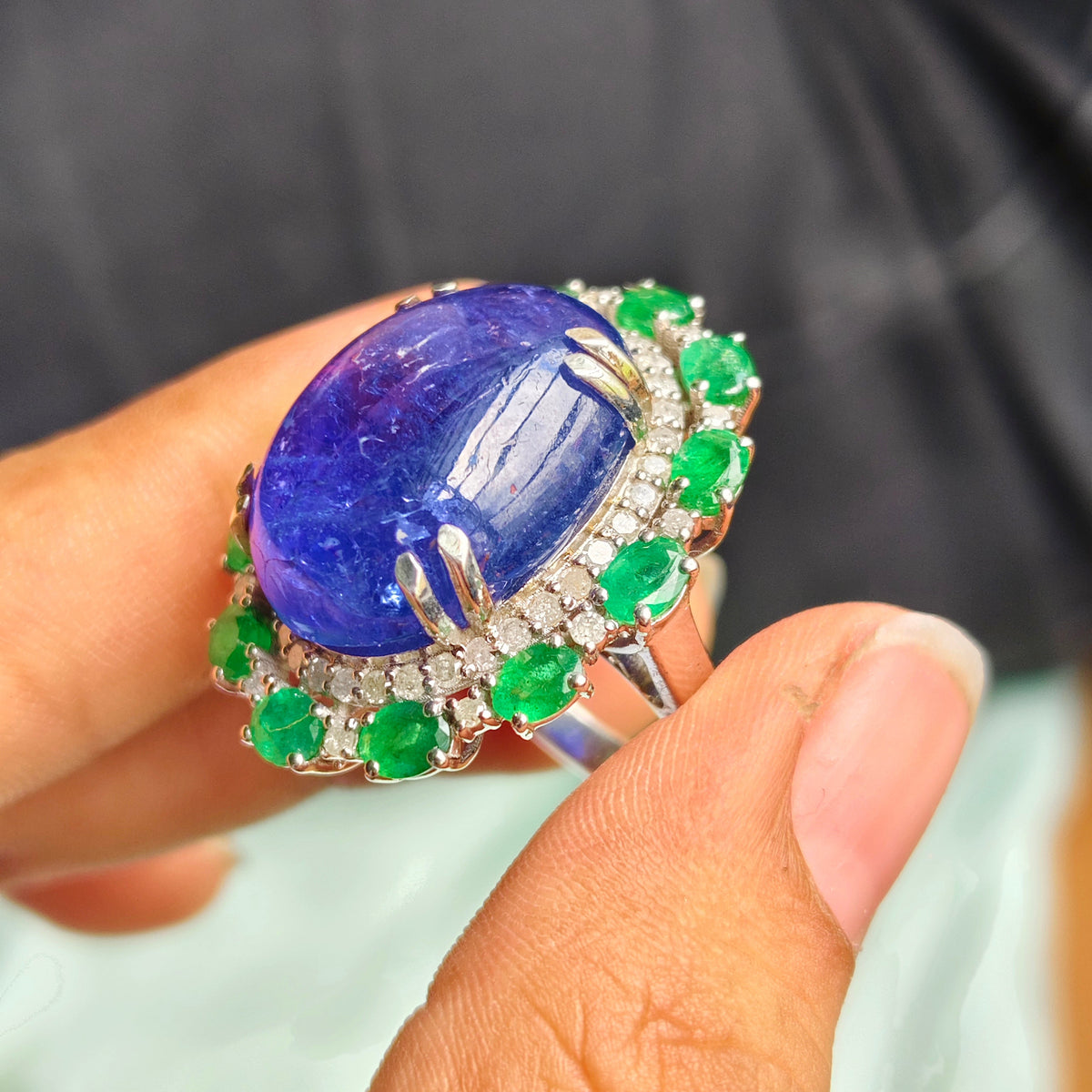 A close-up of a hand holding a large oval ring featuring a deep blue cabochon stone surrounded by a halo of small green gemstones and clear stones on a silver-toned band.