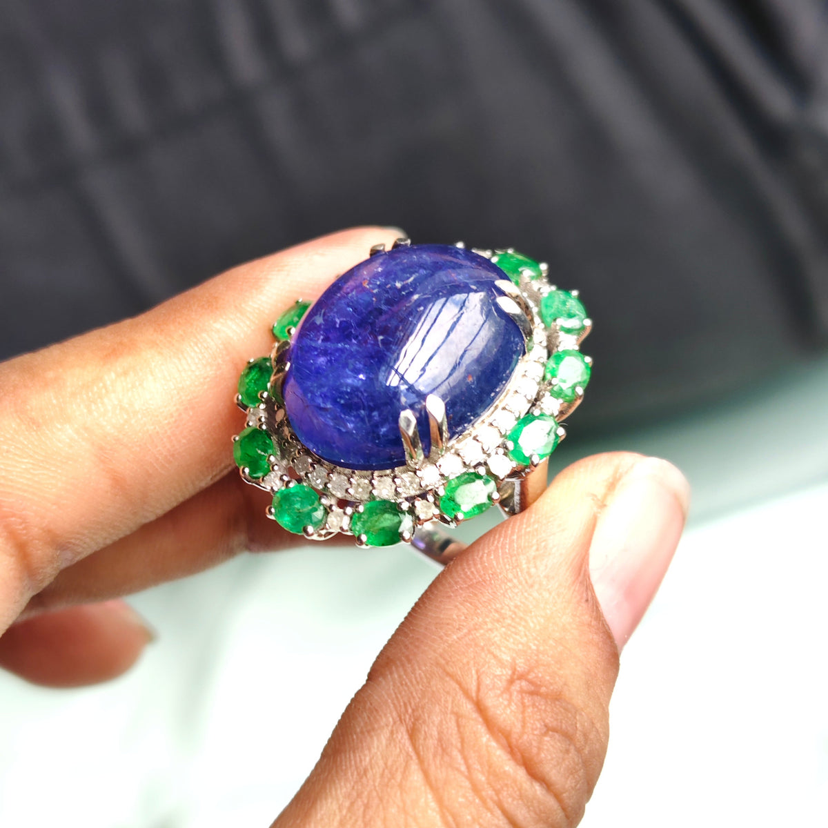 A close-up of a hand holding a large oval ring with a deep blue cabochon center stone and a halo of bright green gemstones in a silver-toned setting.