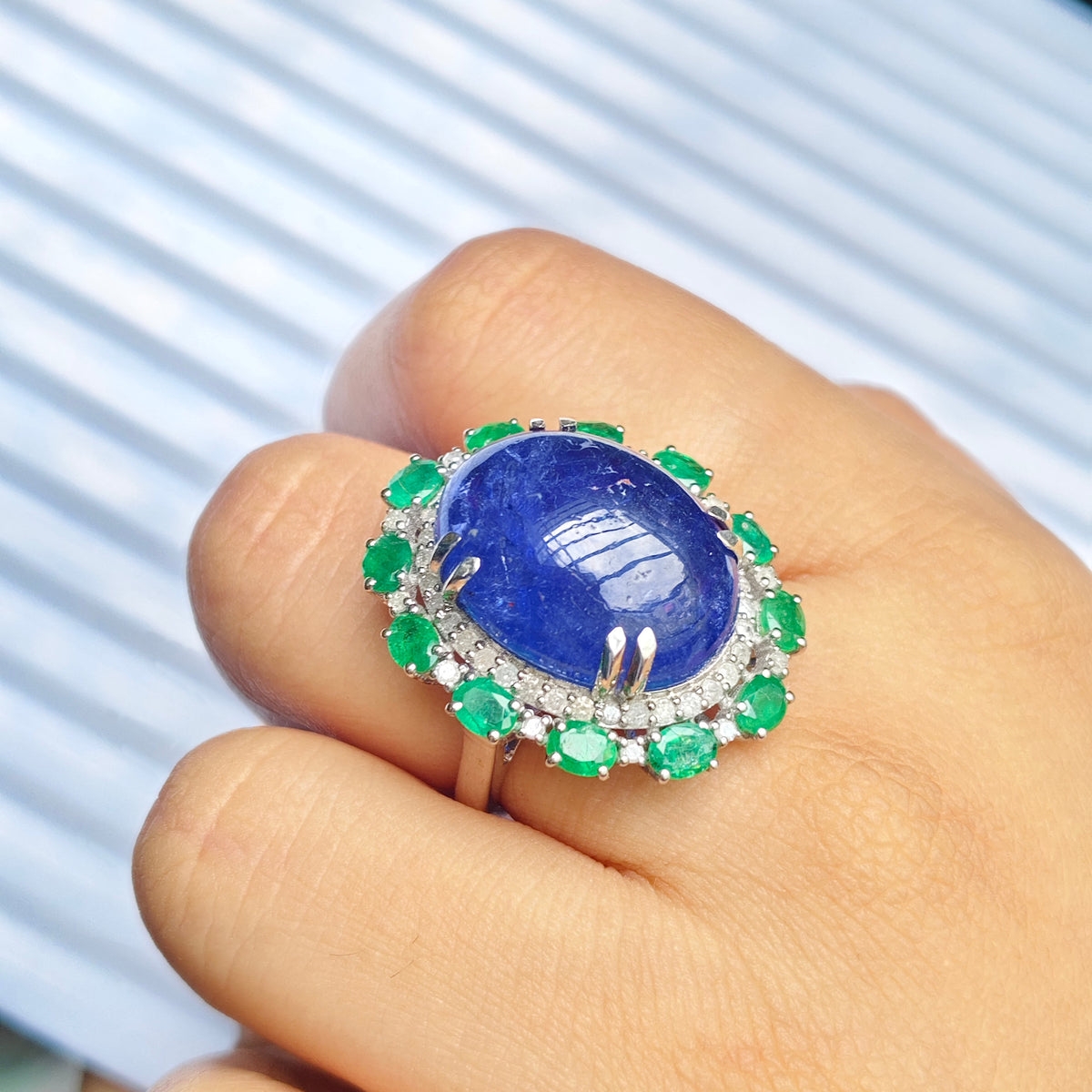 A close-up of a hand wearing a large oval ring featuring a deep blue cabochon stone surrounded by a halo of green emerald-cut stones and small clear stones, set against a blurred striped background.
