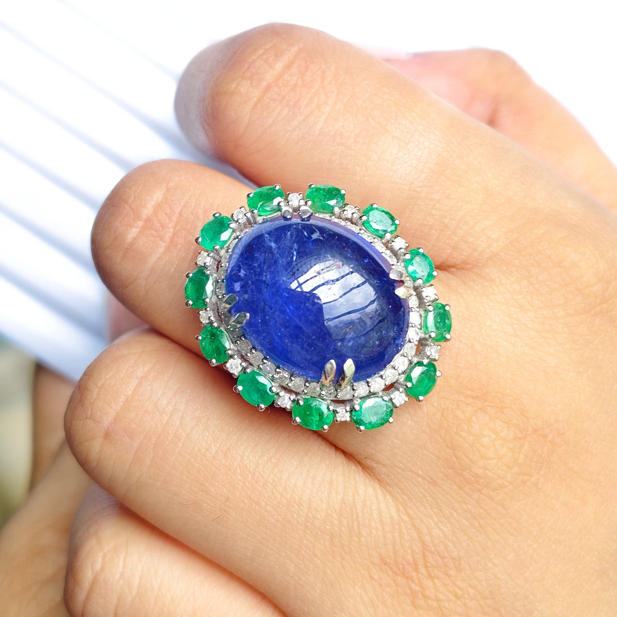 A close-up of a hand wearing a large ring featuring a prominent oval blue lapis lazuli stone surrounded by a halo of green emeralds and small clear stones.