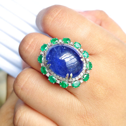A close-up of a hand wearing a large ring featuring a prominent oval blue lapis lazuli stone surrounded by a halo of green emeralds and small clear stones.