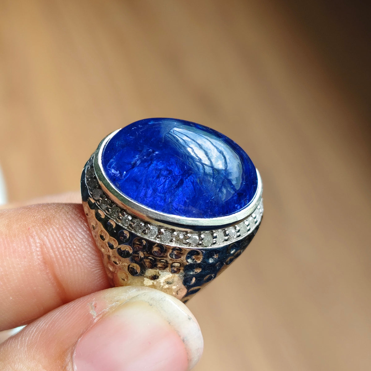 A close-up of a hand holding a large oval blue gemstone ring with a silver-toned textured band and small clear stones around the setting, against a blurred brown background.