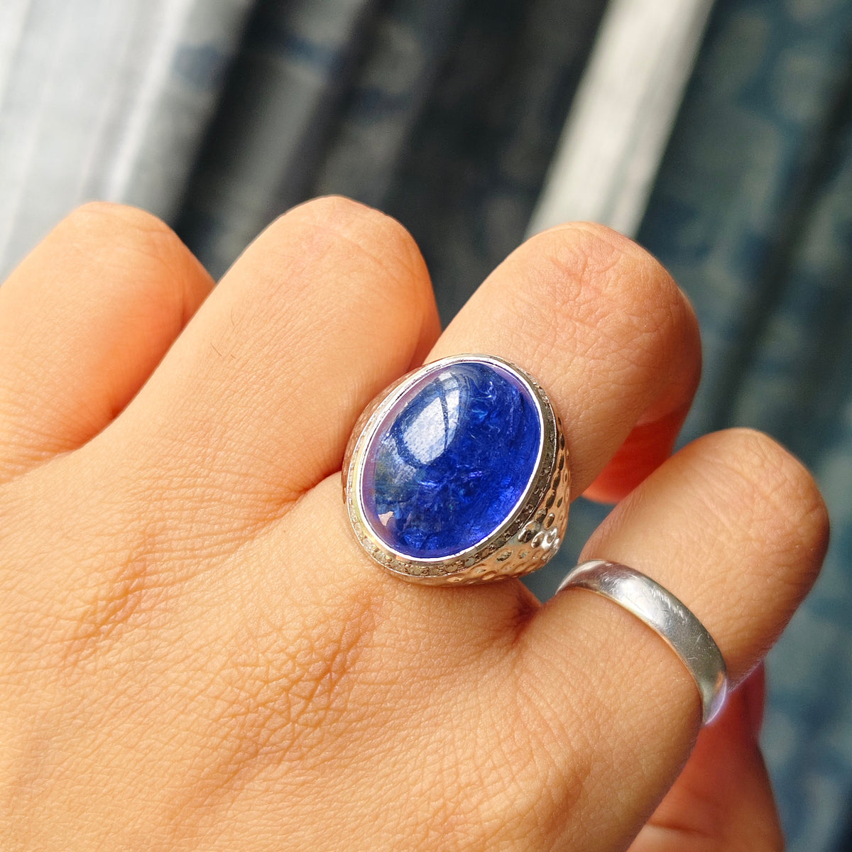 A silver ring with a large oval blue lapis lazuli stone set on a person's finger, with a simple silver band on another finger, against a blurred blue and white background.