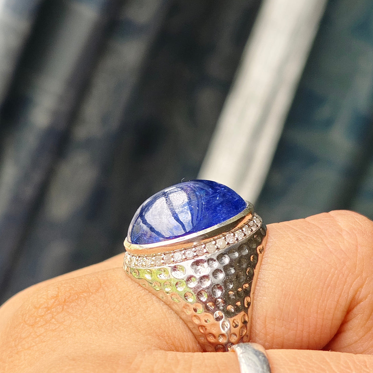 A close-up of a hand wearing a textured silver ring with a polished blue oval gemstone, alongside a simple silver band on another finger, set against a blurred patterned curtain background.