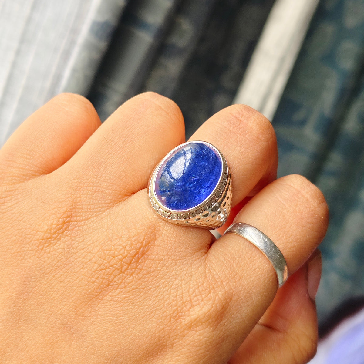 A silver-toned signet ring with a large oval blue lapis lazuli stone, worn on a person's middle finger, with a plain silver band on the ring finger. The background shows a blurred patterned curtain.