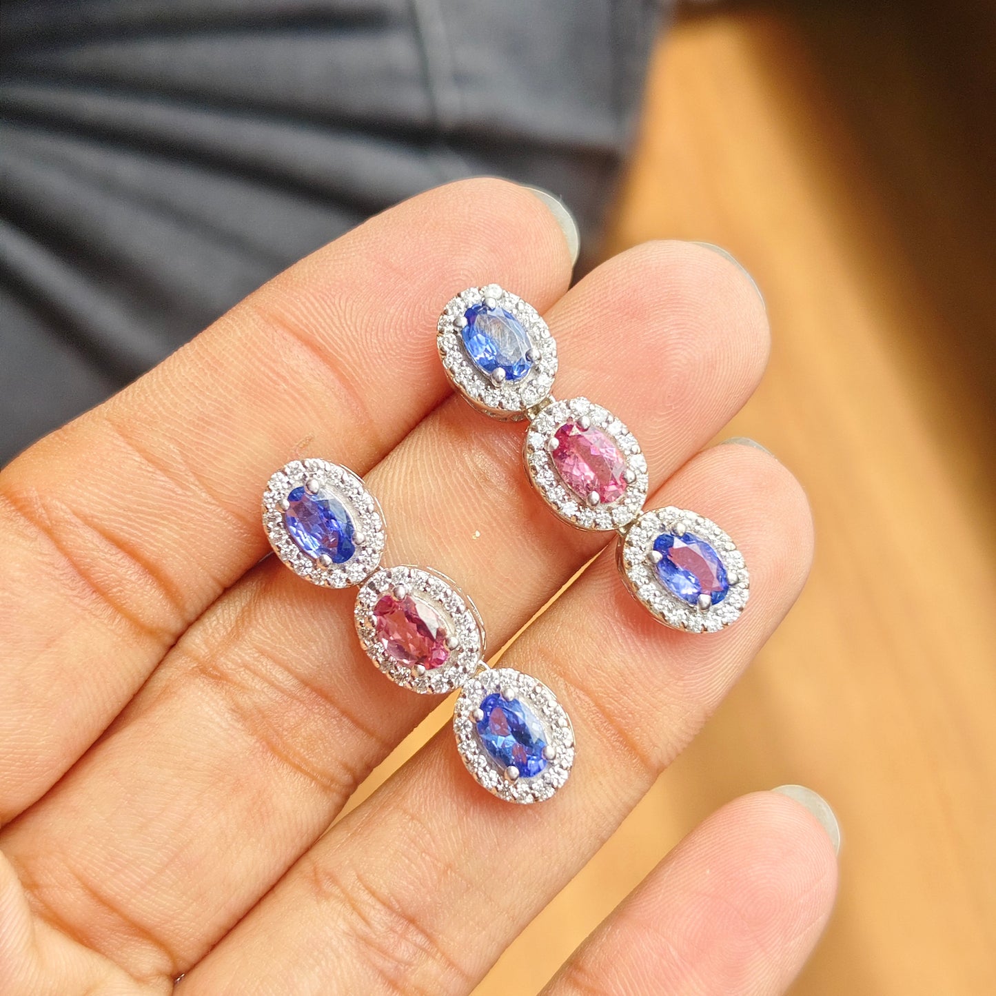A pair of silver-toned drop earrings with oval-cut blue and pink gemstones surrounded by small clear stones, held on a person's fingers against a blurred background.