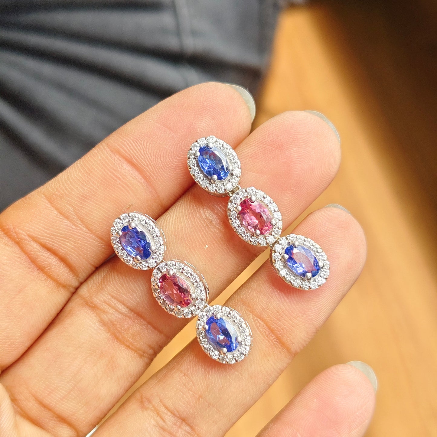 A close-up of a hand holding a pair of silver-toned drop earrings with oval blue and pink gemstones surrounded by small clear stones.