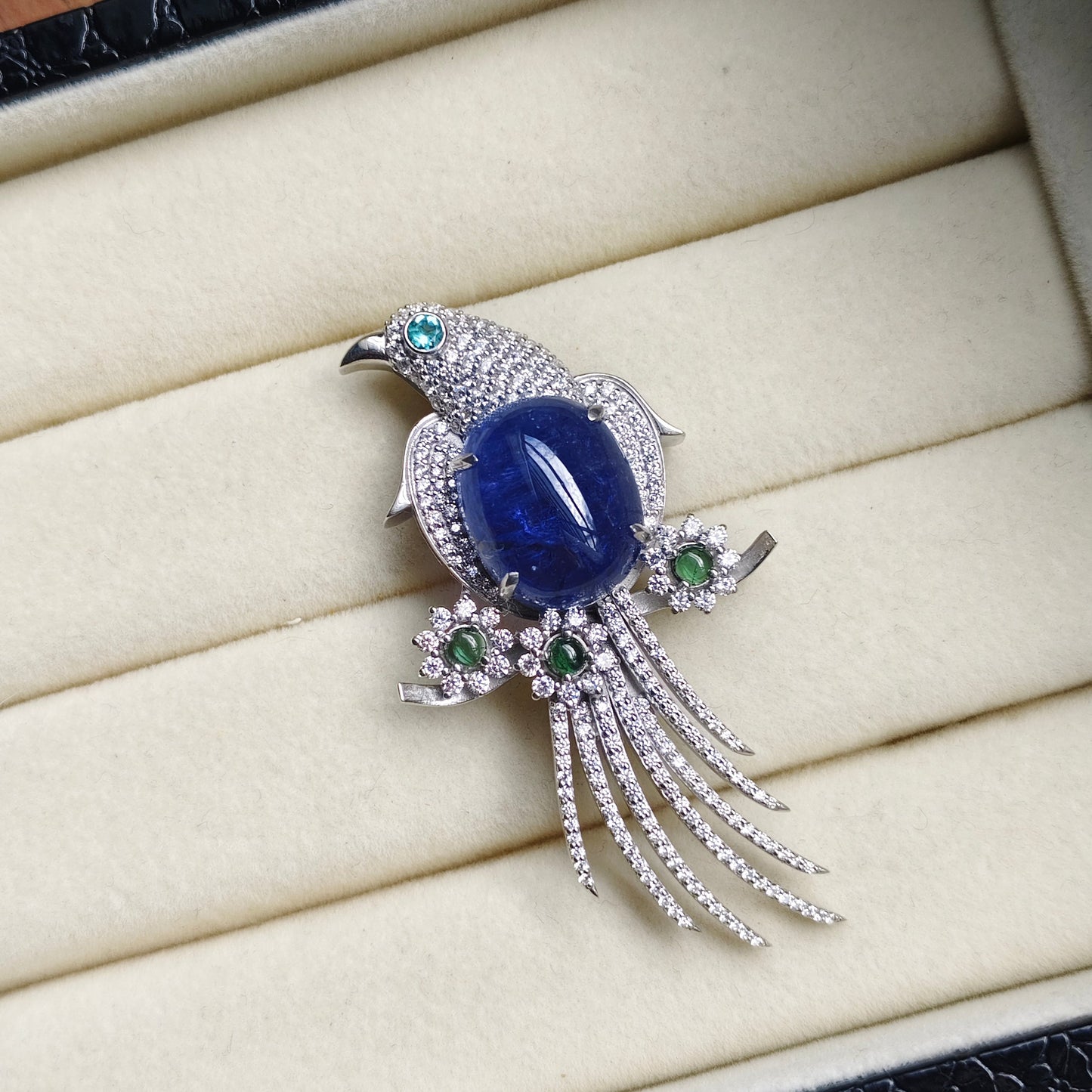 A bird-shaped brooch with a large oval blue gemstone center, silver-toned pavé detailing, and small green accent stones, displayed on a cream jewelry box.