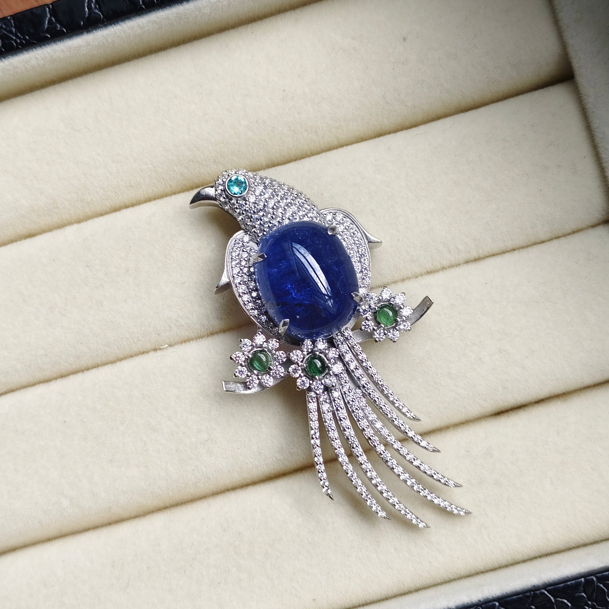 A bird-shaped brooch with a large oval blue gemstone center, silver-toned pavé detailing, and small green accent stones, displayed on a cream jewelry box.