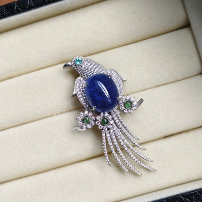 A bird-shaped brooch with a large oval blue gemstone center, silver-toned pavé detailing, and small green accent stones, displayed on a cream jewelry box.