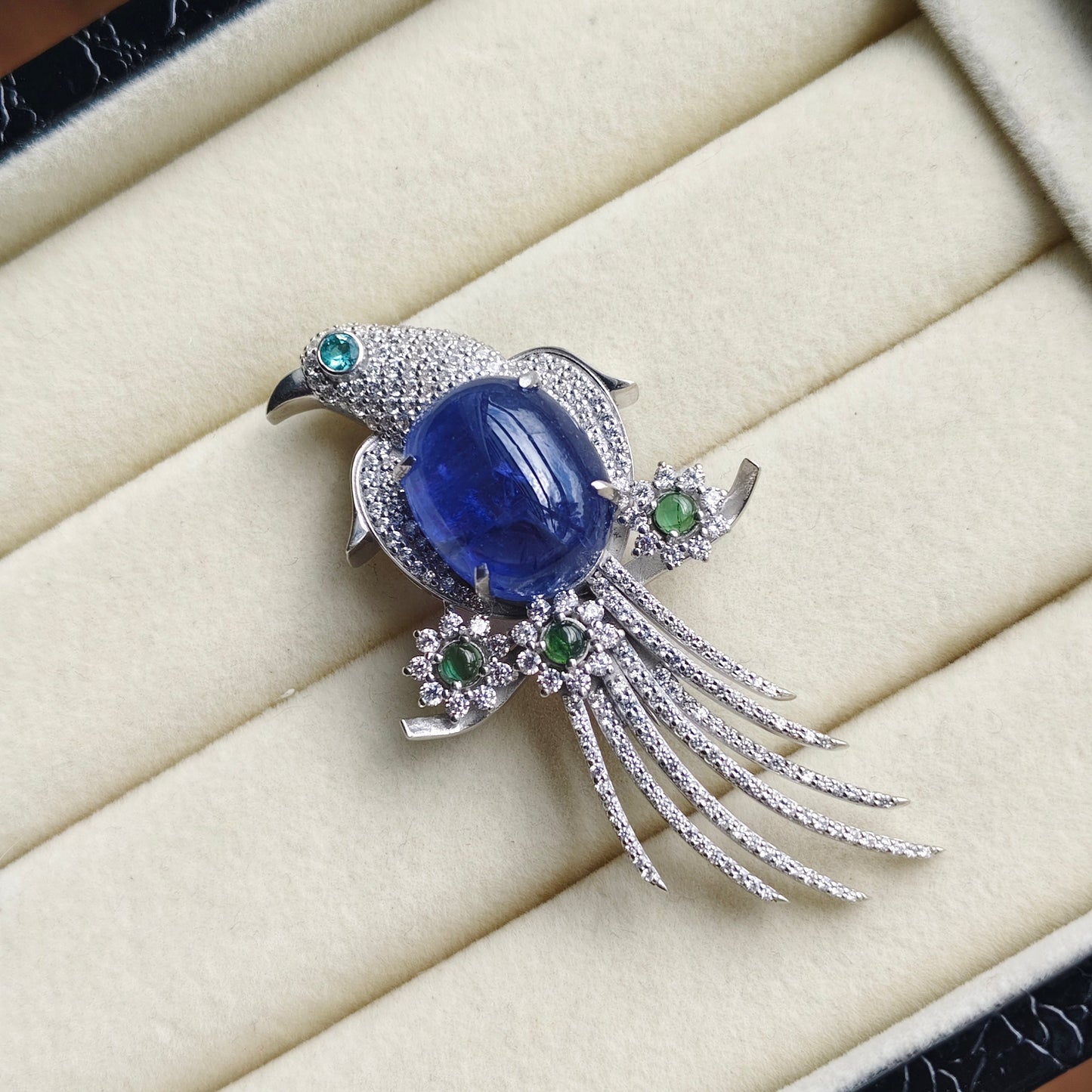 A bird-shaped brooch with a large oval blue gemstone body, silver-toned pavé setting, and small green gemstone accents, displayed on a cream jewelry box cushion.
