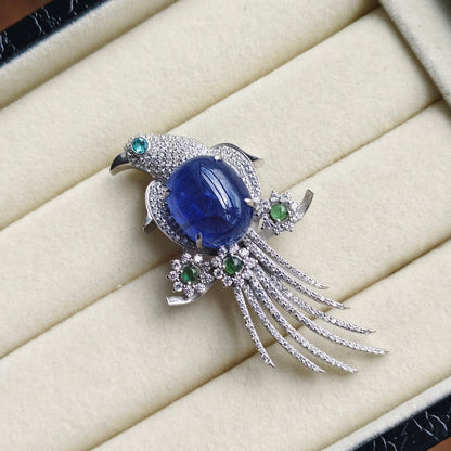 A bird-shaped brooch with a large oval blue gemstone body, silver-toned pavé setting, and small green gemstone accents, displayed on a cream jewelry box cushion.