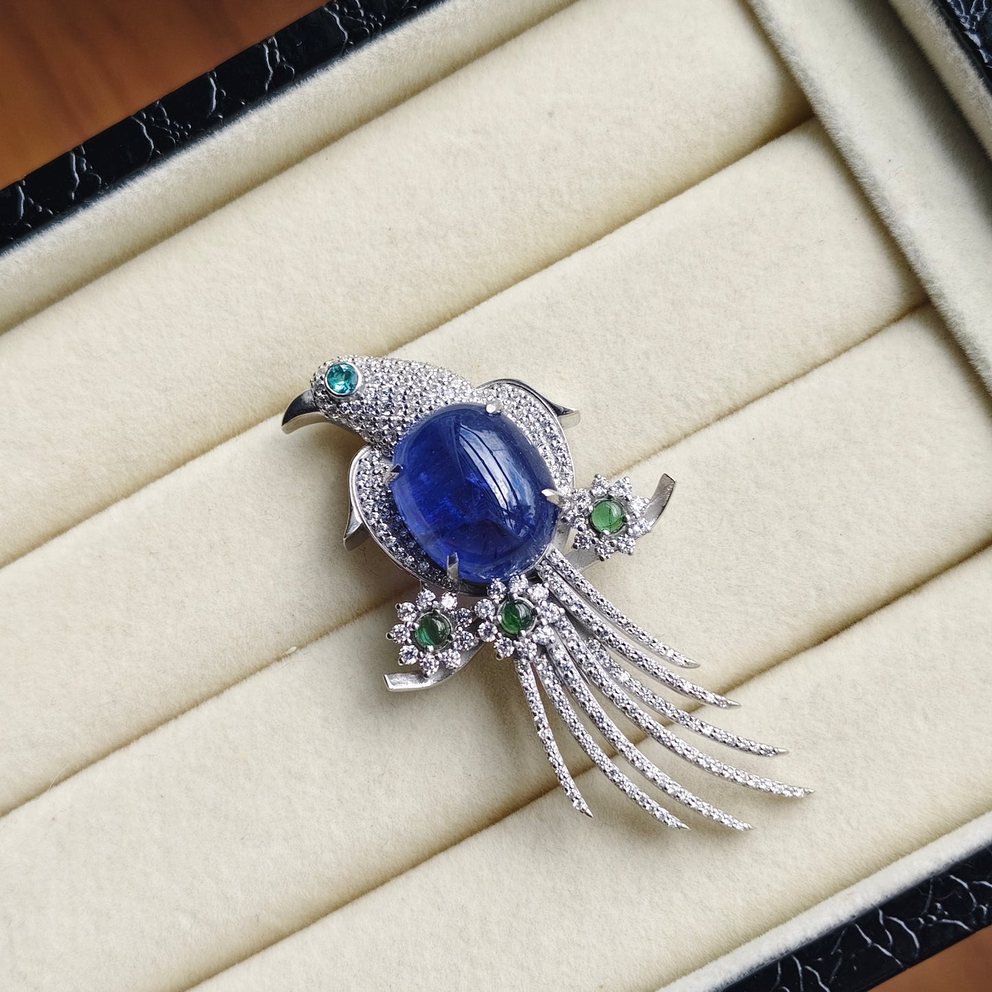 A bird-shaped brooch with a large oval blue cabochon center stone, silver-toned pavé detailing, and small green gemstone accents, displayed on a cream jewelry box interior.