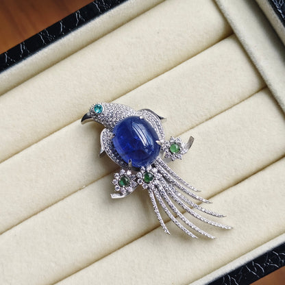 A bird-shaped brooch with a large oval blue cabochon center stone, silver-toned pavé detailing, and small green gemstone accents, displayed on a cream jewelry box interior.