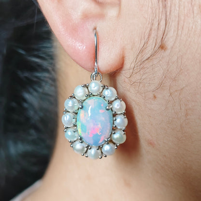 A close-up of a person's ear wearing a dangling earring with a silver-tone hook and a round opal-like center stone surrounded by small white faux pearls.