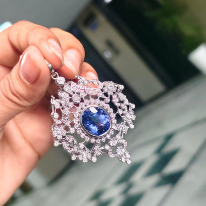 A hand holding a silver filigree pendant featuring a central oval blue gemstone surrounded by small clear stones, with a blurred indoor background.