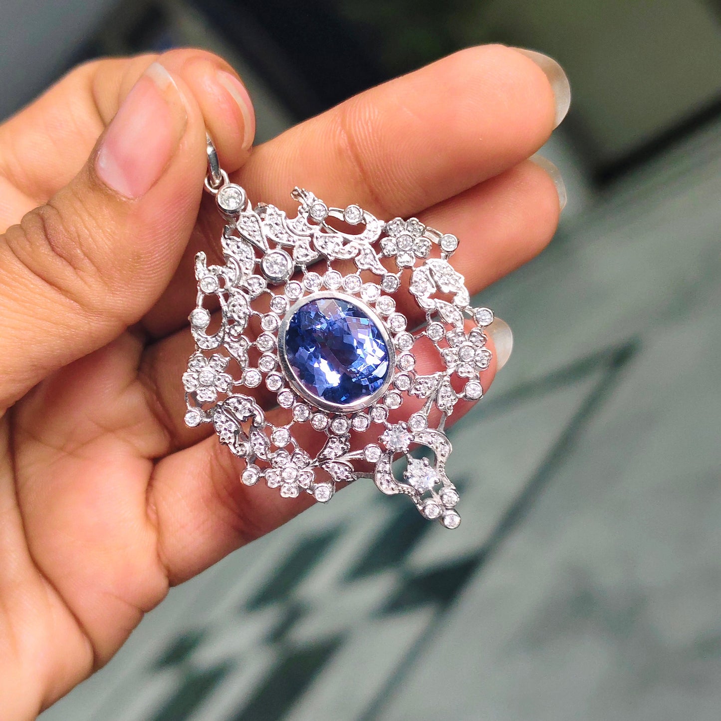 A hand holding a silver-toned pendant featuring a central oval blue sapphire surrounded by intricate pavé clear stones, with a blurred indoor background.