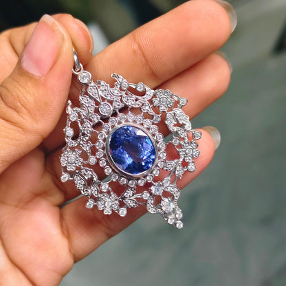 A hand holding a silver filigree pendant featuring a central oval blue gemstone surrounded by clear stones.