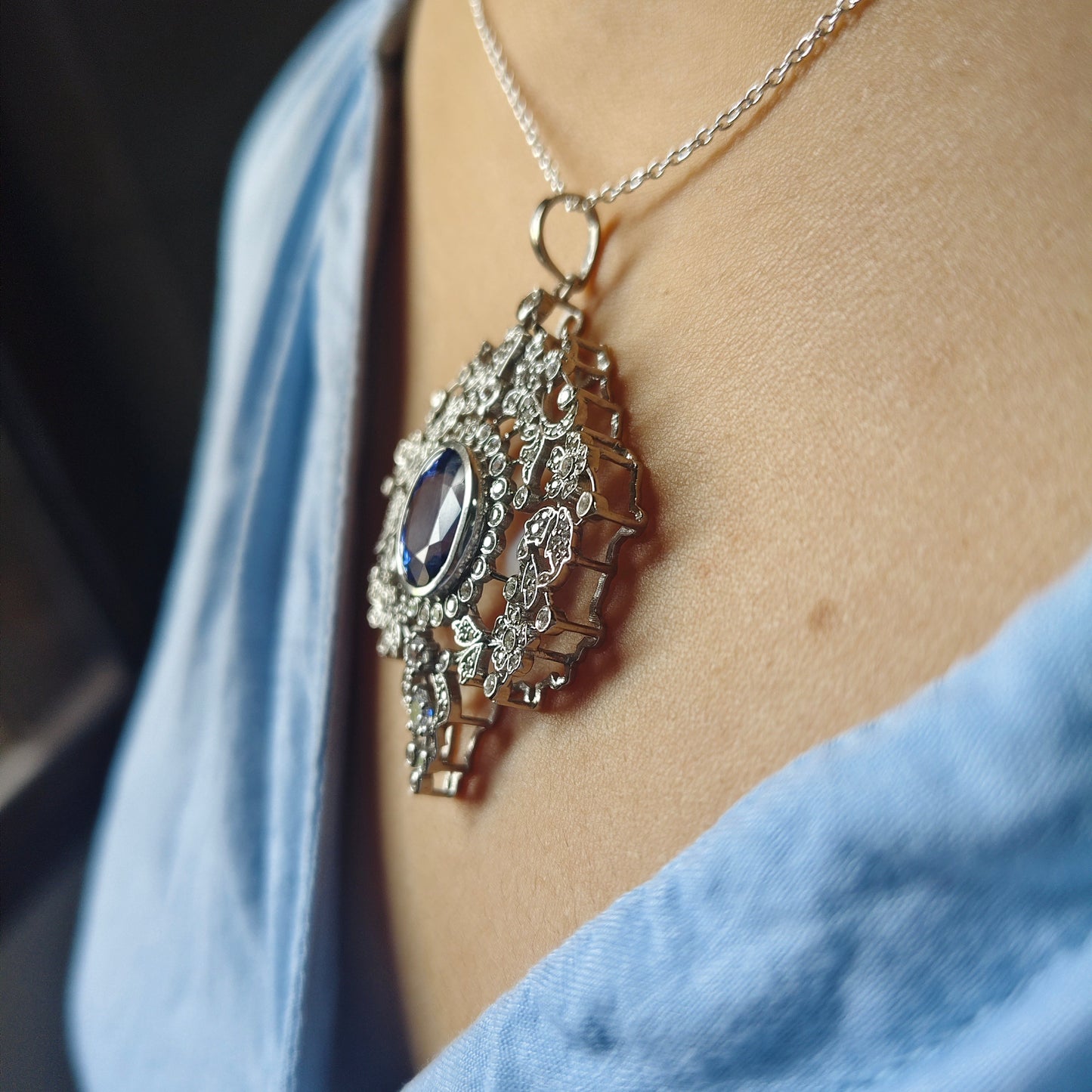 A close-up lifestyle shot of a woman wearing a detailed silver pendant necklace with a central dark blue oval stone, styled with a light blue shirt.
