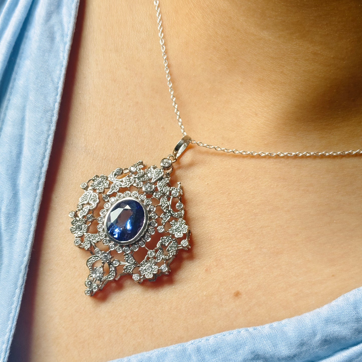 A close-up of a silver pendant necklace with a central oval blue gemstone and surrounding clear stones, worn on a model's neck over a light blue top.