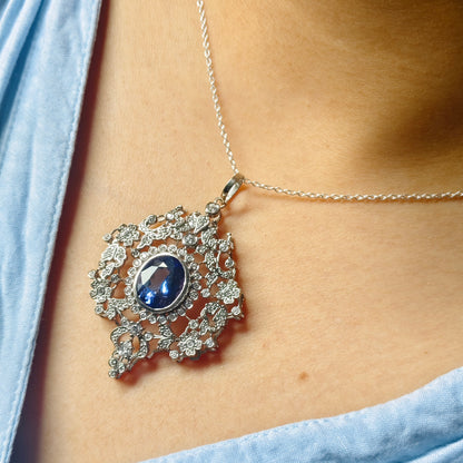A close-up of a silver pendant necklace with a central oval blue gemstone and surrounding clear stones, worn on a model's neck over a light blue top.