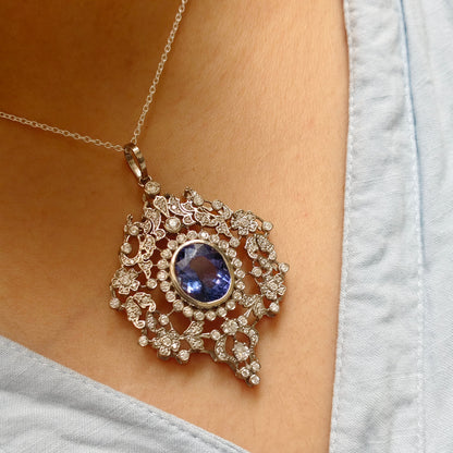 A close-up of a person wearing a silver-toned pendant necklace with a large oval blue gemstone surrounded by intricate floral crystal detailing, paired with a light blue denim top.