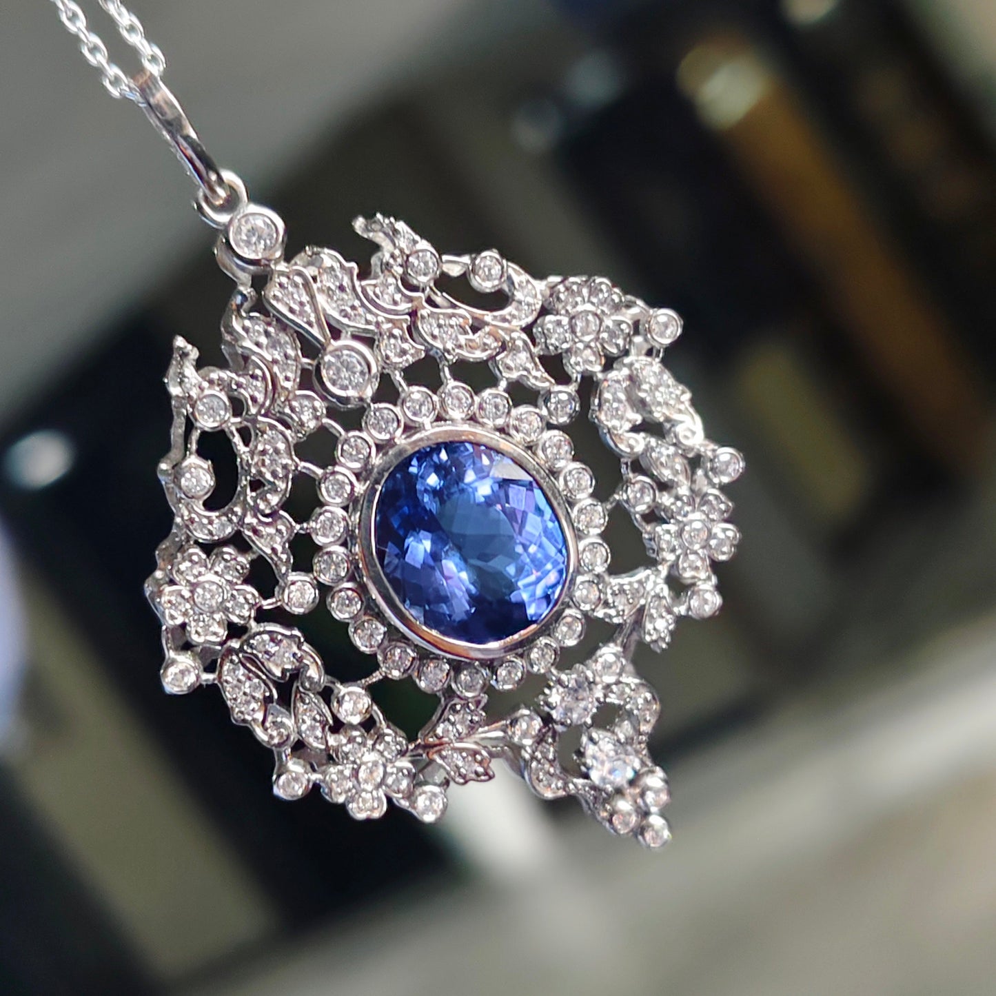 An ornate silver-toned pendant necklace featuring a central oval blue gemstone surrounded by intricate floral and scroll motifs set with small clear stones, hanging from a delicate chain against a blurred indoor background.